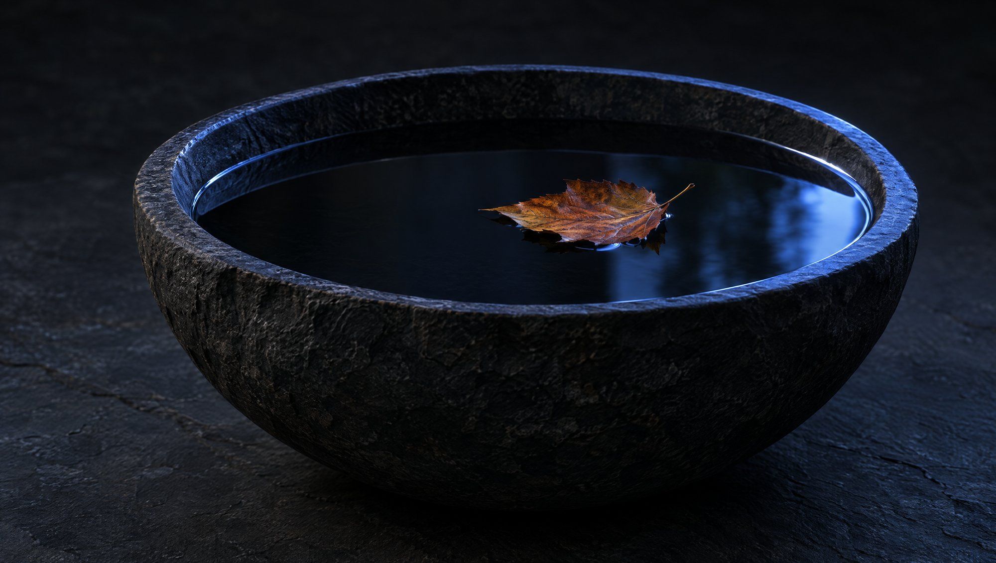 A dark, rough stone bowl filled with still black water on a slate surface, with a single brown fallen leaf floating on the water and a thin rim of indigo light along the bowl's edge.
