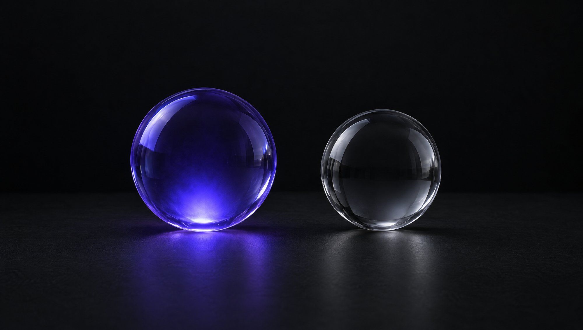 Two clear glass spheres side by side on a dark surface, the larger one glowing indigo from inside and the smaller one lit only in neutral grey.